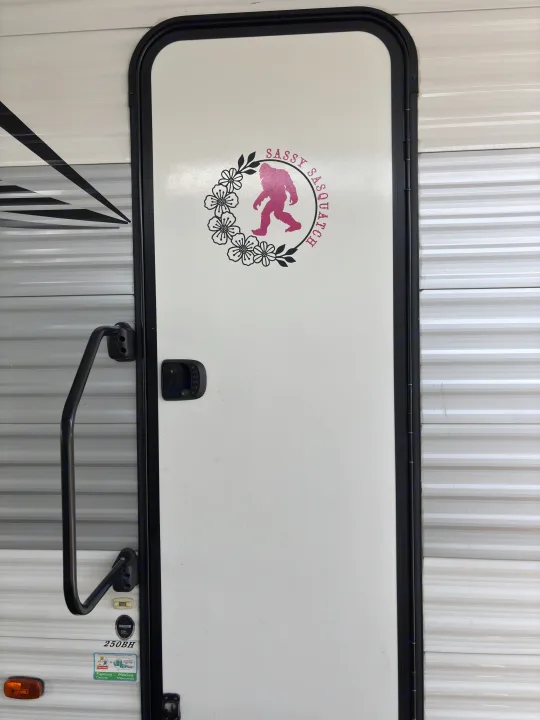 White RV rental door with a pink 'Sassy Sasquatch' decal in a floral wreath, showing a grab handle.. East to West Della Terra 2021