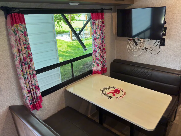Bright RV rental dinette featuring a 'Sassy Sasquatch' table, floral curtains, window view to nature, and mounted TV.. East to West Della Terra 2021