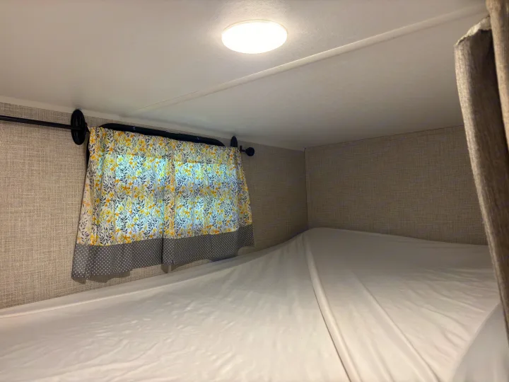 An RV rental's cozy bunk bed, complete with a bright floral curtain and textured walls, offers comfort for travel.. East to West Della Terra 2021