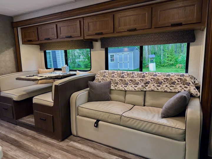 Spacious RV rental living area with dinette, sofa, and storage cabinets, offering views of a wooded yard.. Forest River Sunseeker LE 2021