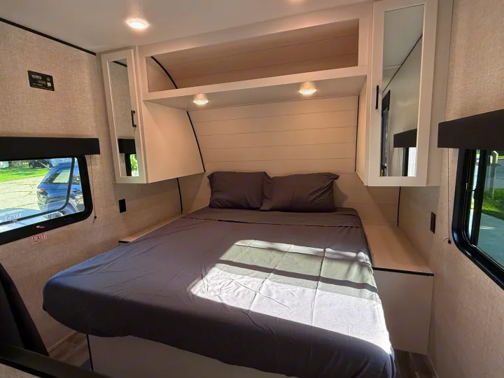 This bright RV rental bedroom features a queen bed with gray linens, overhead storage, mirrored cabinets, and side windows.. Jayco Jay Flight 2025