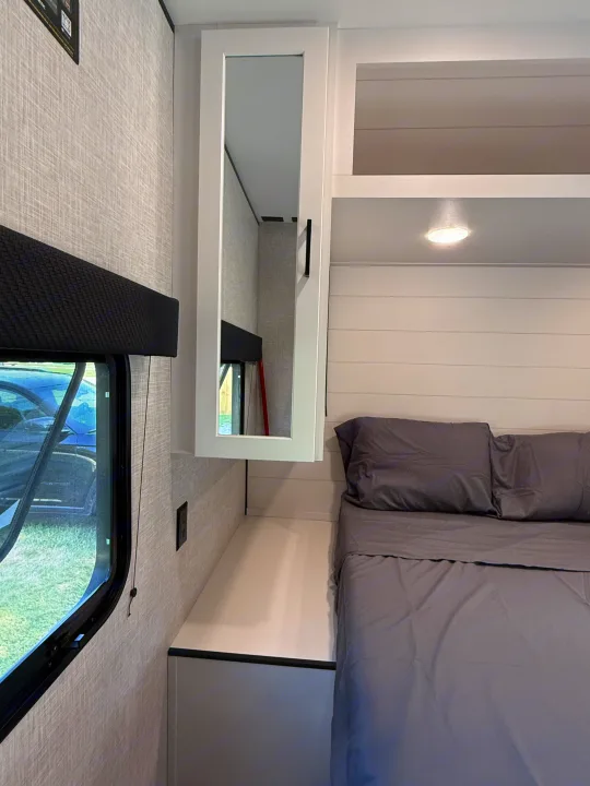Compact RV rental bedroom showing a gray bed, mirrored cabinet, window, and wall outlet. Cozy and functional space.. Jayco Jay Flight 2025