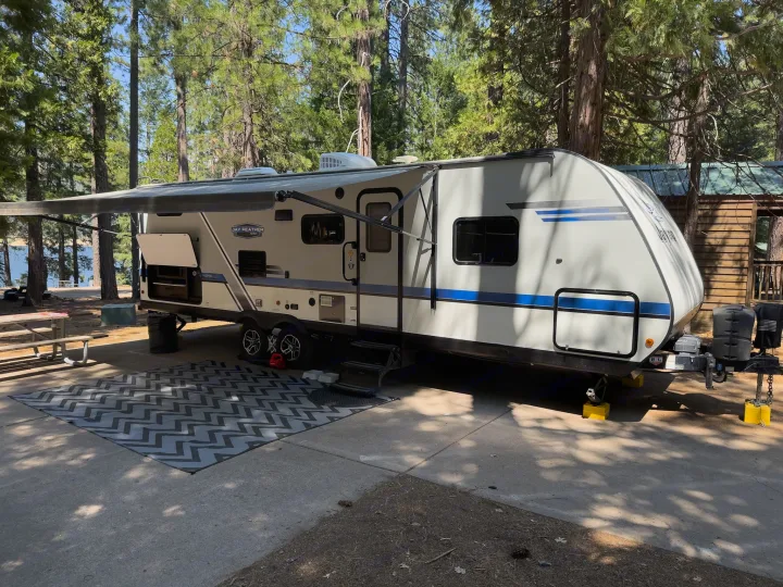 Travel trailer RV rental at a sunny forest campsite. Awning extended over a mat, offering outdoor comfort for nature trips.. Jayco Jay Feather 2019