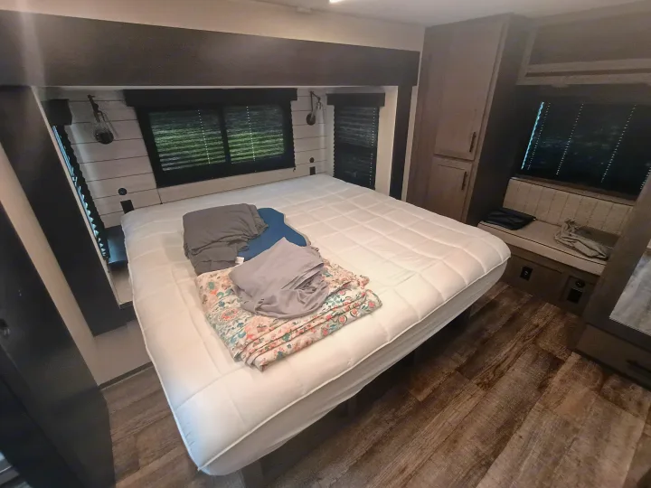 An RV rental bedroom with a white mattress, folded linens, window blinds, and built-in wood-look cabinets. Modern & functional.. Forest River Arctic Wolf 2025