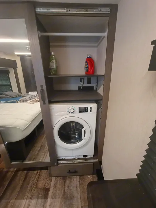 Inside an RV rental, a built-in closet houses a compact washing machine and laundry supplies.. Forest River Arctic Wolf 2025