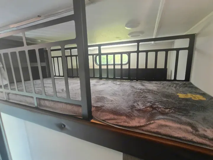 Top bunk bed in an RV rental, featuring a soft blanket and safety railing. A window is visible in the background.. Forest River Arctic Wolf 2025