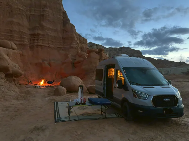 An RV rental camper van is parked amidst red rock formations at dusk, with a campfire and chairs set up for cozy desert camping.. Ford Transit 2022