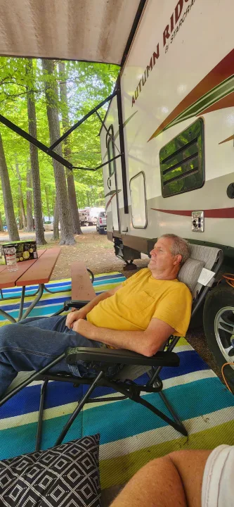 Man relaxing in a chair under an RV rental's awning at a wooded campground, beside a picnic table. Peaceful outdoor scene.. Starcraft Autumn Ridge 2014