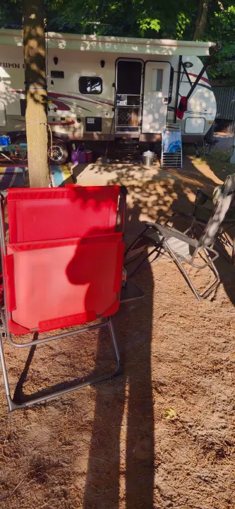Sunny RV rental campsite scene with red chair, other camping chairs, and an RV under shade trees.. Starcraft Autumn Ridge 2014