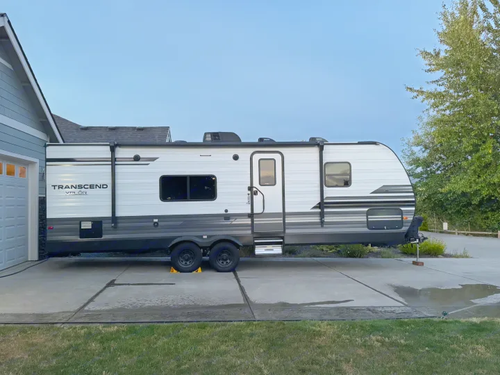 This Transcend Valor rv rental, a white & gray travel trailer, is parked on a driveway next to a house.. Grand Design Transcend Xplor 2021