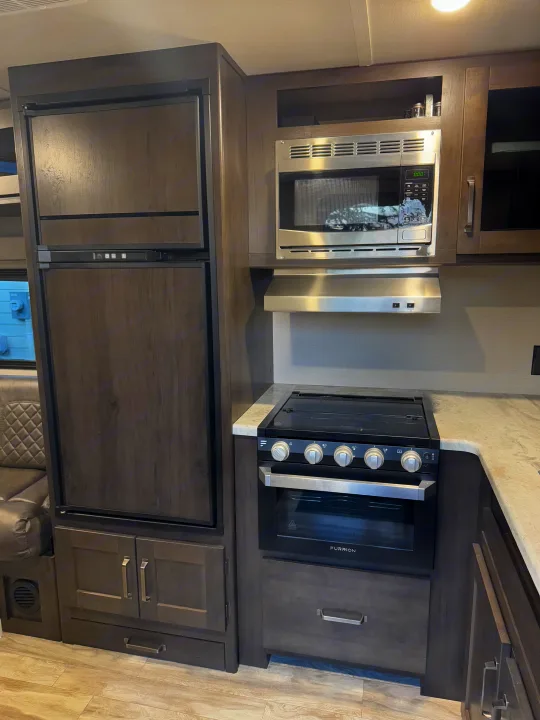 A well-equipped RV rental kitchen with dark wood-grain cabinets, fridge, microwave, and a Furrion stove/oven.. Grand Design Transcend Xplor 2021