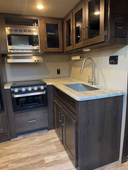 A spacious RV rental kitchen with stainless steel microwave, oven, sink, dark cabinets, and light stone-look counters.. Grand Design Transcend Xplor 2021