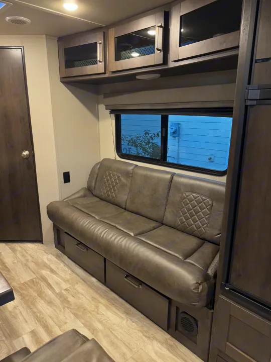 Inside this RV rental: a gray sofa with storage, overhead cabinets, a window, and wood-look flooring.. Grand Design Transcend Xplor 2021