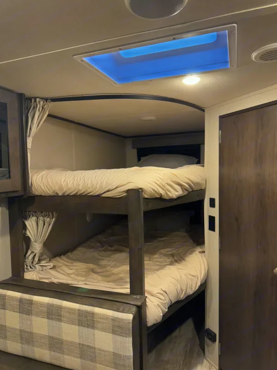 Inside an RV rental, showing comfortable bunk beds with clean bedding, a skylight, and compact design.. Grand Design Transcend Xplor 2021