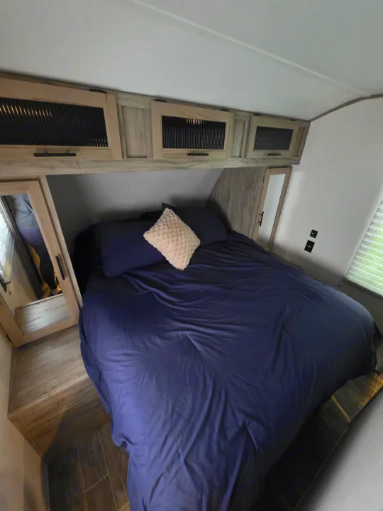 Cozy RV rental bedroom: large bed with navy linens, light pillow, and smart overhead storage. Person reflected in mirror.. Forest River Cherokee Alpha Wolf 2021