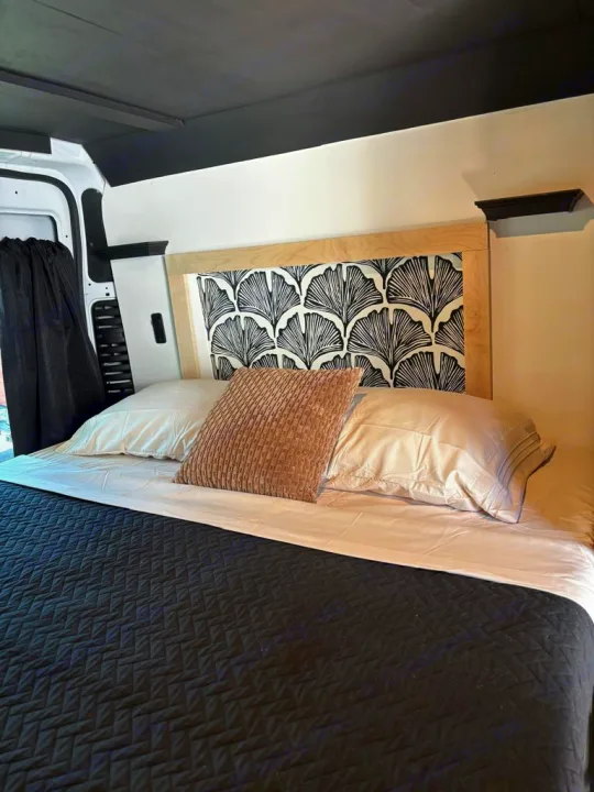 Cozy RV rental interior featuring a bed with patterned headboard, plush pillows, and a black quilted blanket.. Ram Promaster 2022