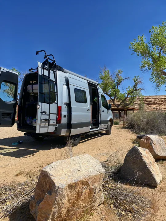 Silver RV rental van with open doors at a sunny desert campsite, surrounded by rocks and trees. Adventure awaits.. Winnebago Revel 2020