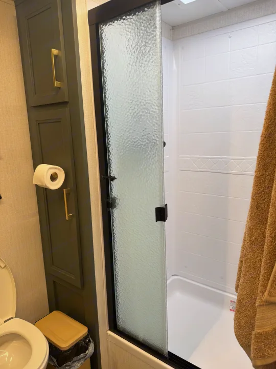 RV rental bathroom featuring a compact toilet, dark cabinets, and a textured glass shower door.. Heartland RVs Eddie Bauer 2024