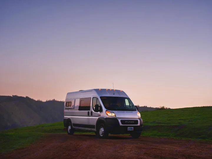 A silver camper van on a dirt road, headlights on, with green hills and sunset sky. Ideal for your next RV rental adventure.. Wayfarer Walt 2021