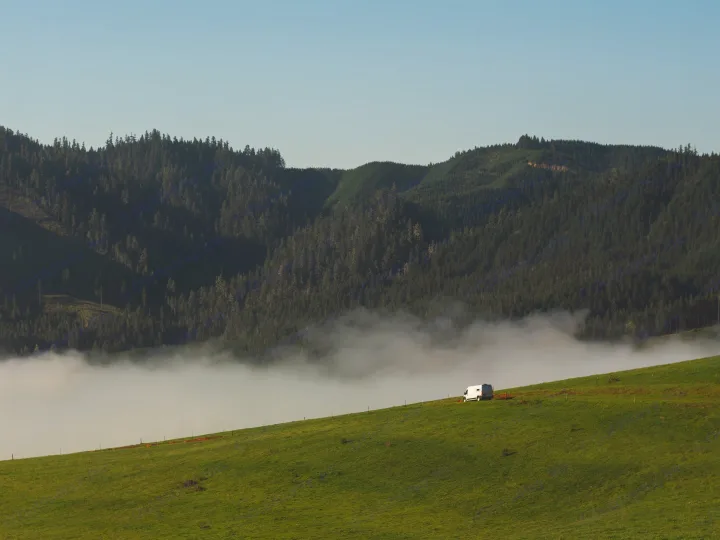 White rv rental on a green hill, with low fog blanketing the valley and dense forests beyond.. Wayfarer Walt 2021