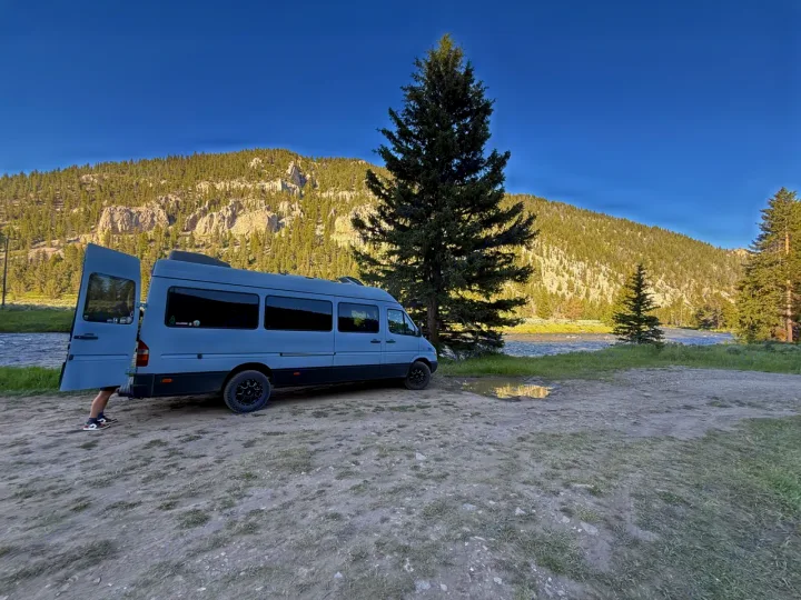 Grey RV rental camper van by a river & mountains. Rear doors open, showing legs. Perfect outdoor travel.. Dodge Sprinter 2006