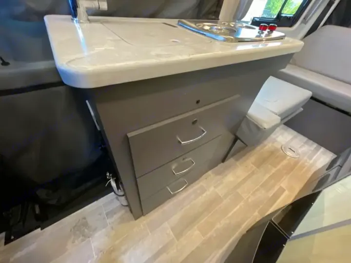 Compact kitchenette in an RV rental, featuring a counter, sink, stove, and drawers, alongside a comfy side bench.. Thor Tranquility 2022