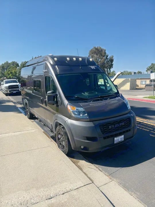 A dark gray Ram ProMaster camper van with rooftop lights, ideal for an rv rental, parked on a sunny street.. Winnebago Solis 2022