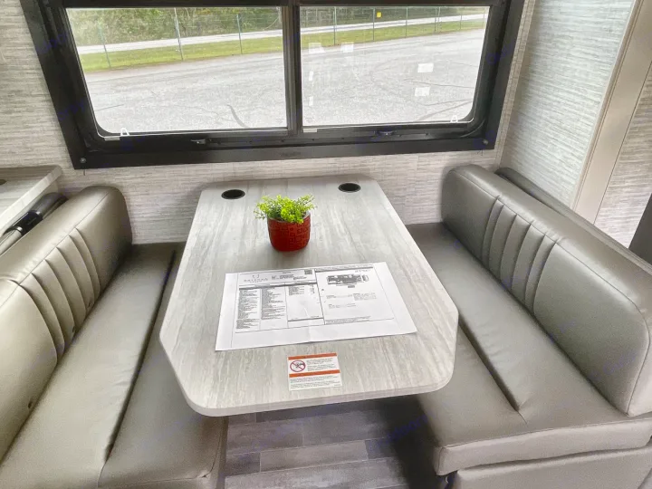 An RV rental's dinette area: grey upholstered booth seats, a light wood table with a plant & papers, and a large window view.. Entegra Odyssey 2025