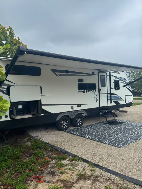 White Keystone Passport RV rental travel trailer set up at a campsite with awning, outdoor kitchen, and ground mat.. Keystone RV Passport TT 2022