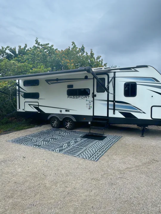 An RV rental at a gravel campsite, featuring an extended awning & decorative outdoor rug.. Keystone RV Passport TT 2022