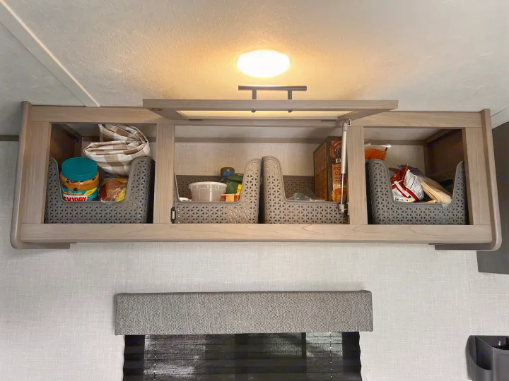 Organized RV rental overhead cabinet: interior view showing gray storage bins with pantry items like snacks and peanut butter.. Dutchmen Coleman Lantern LT 2023