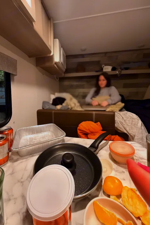Inside an rv rental: a cozy kitchen area with pans, food items, and a person relaxing on the dinette in the background.. Dutchmen Coleman Lantern LT 2023