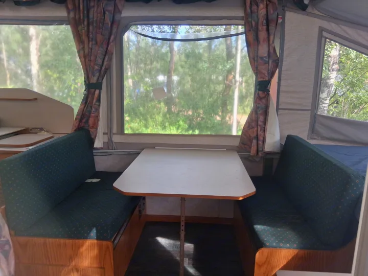 Inside an RV rental pop-up camper: a cozy dinette with patterned benches and a table, overlooking nature through clear windows.. Bonair 1050C 1996
