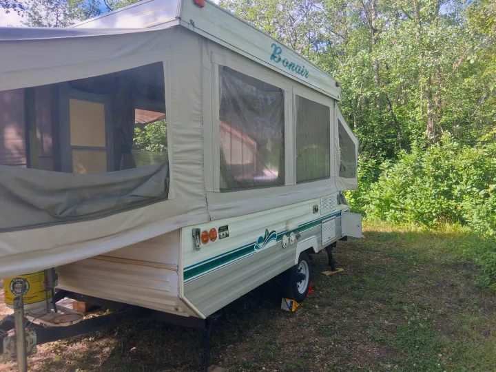 A light-colored Bonair pop-up camper, ideal for rv rental, is set up in a sunny, green forest campsite.. Bonair 1050C 1996