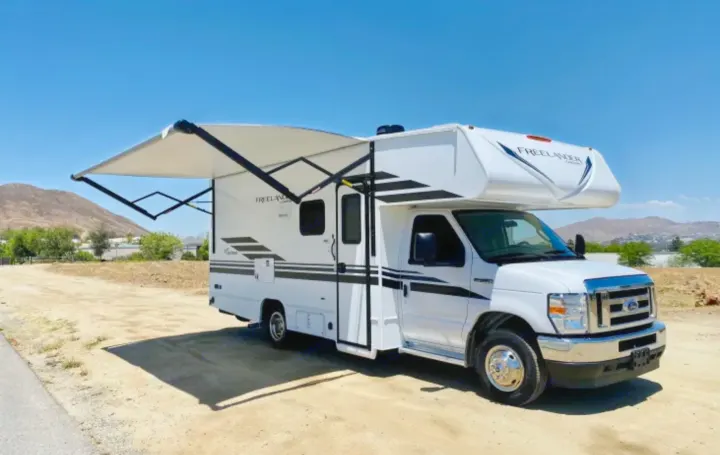 A white Class C RV rental, with its awning extended, is parked on a dirt lot under a bright blue sky, mountains visible.. Forest River Coachmen Freelander 2021