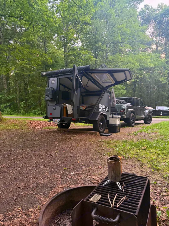 Explore the wild! This rv rental setup features a Tiger Moth overland trailer & Jeep at a forested campsite with a fire pit.. TAXA TAXA Trailer 2019