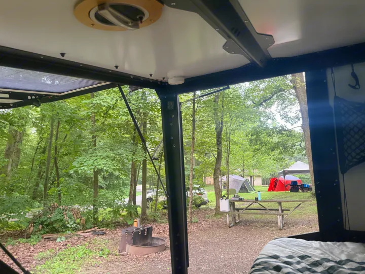 Relaxing inside an RV rental, looking out at a peaceful forest campsite with tents, a picnic table, and a fire pit.. TAXA TAXA Trailer 2019