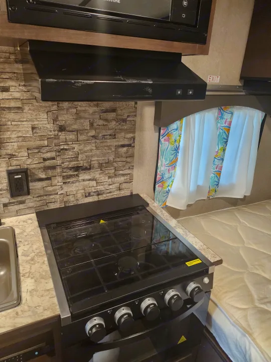 RV rental kitchen: Compact space featuring a modern stove, sink, and microwave above a tiled backsplash. Ideal for camper travel.. Sunset Park RV Sun Lite 2022