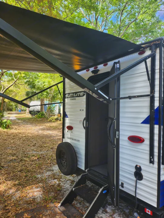 This RV rental travel trailer has its awning extended, with visible entry steps and a spare tire, parked in a sunny campsite.. Sunset Park RV Sun Lite 2022