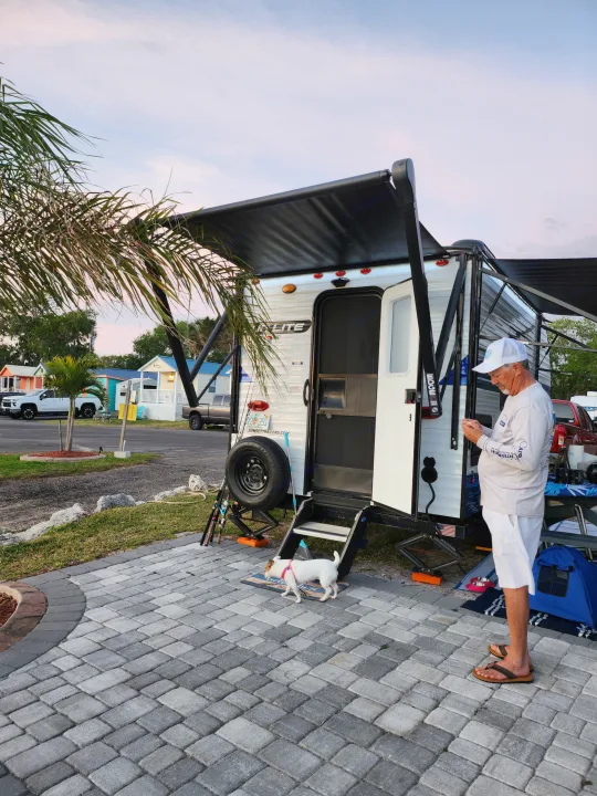 Evening at an RV rental site: Man with dog next to a white travel trailer, awning extended. Colorful homes in background.. Sunset Park RV Sun Lite 2022