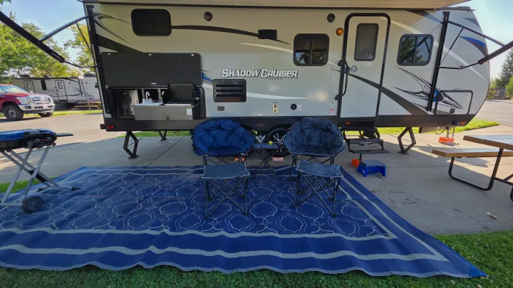 Relaxing by an RV rental with an outdoor kitchen, BBQ grill, comfy chairs, and a large blue patio rug at the campsite.. Shadow Cruiser 240RBH 2019