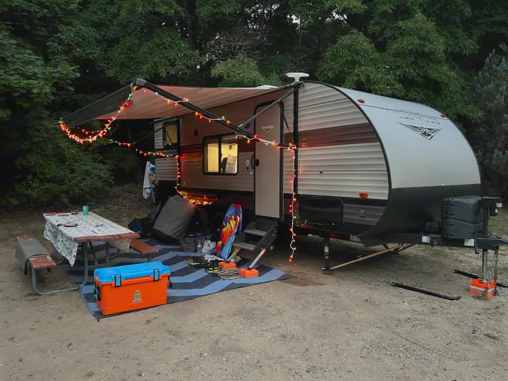 Enjoy this cozy rv rental setup at a shaded campsite. The camper has festive string lights, a picnic table, and cooler for outdoor fun.. Forest River Wildwood X-Lite 2019