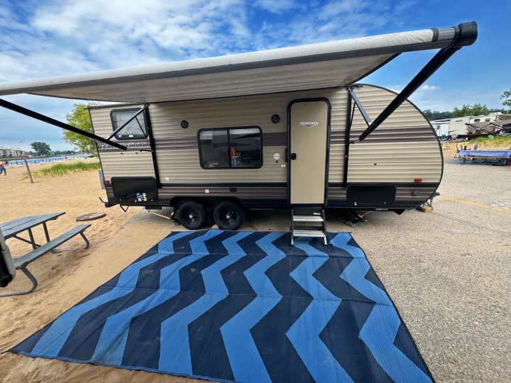 This RV rental is set up at a lakeside campsite with an extended awning and outdoor mat on the sand and pavement.. Forest River Wildwood X-Lite 2019