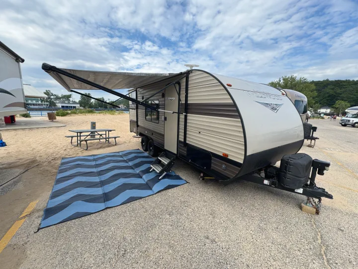 A tan RV rental is parked with an extended awning and a blue/black mat on sand next to a picnic table.. Forest River Wildwood X-Lite 2019