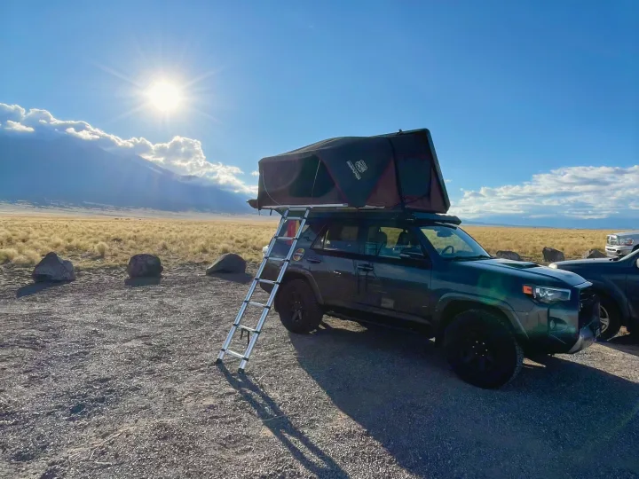 An RV rental SUV with a rooftop tent & ladder set up for camping, with sunny mountains & golden fields in the background.. Toyota 4Runner TRD OffRoad 2020