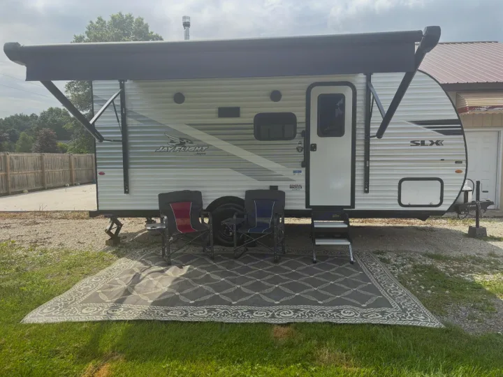 Side view of a white Jay Flight SLX RV rental, parked with an extended black awning. Two camping chairs and an outdoor rug are set up.. Jayco Jay Flight SLX 2025