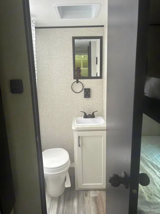View into a compact RV rental bathroom: toilet, sink, mirror, and glimpse of sleeping area.. Jayco Jay Flight SLX 2025
