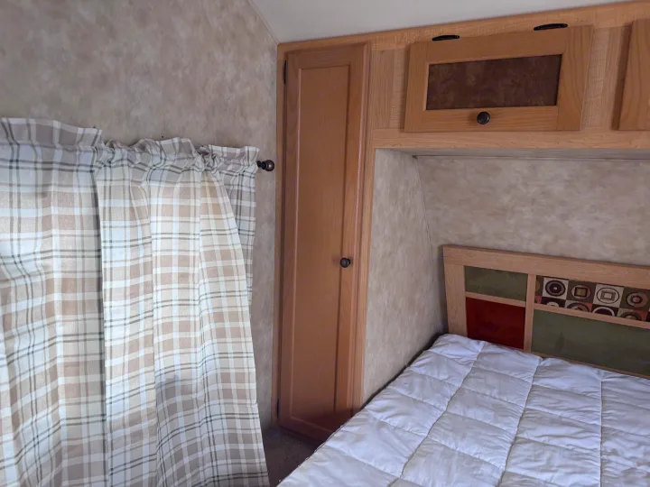 Cozy RV rental bedroom featuring plaid curtains, a bed with a patterned headboard, and wood cabinets.. kz Coyote 2010