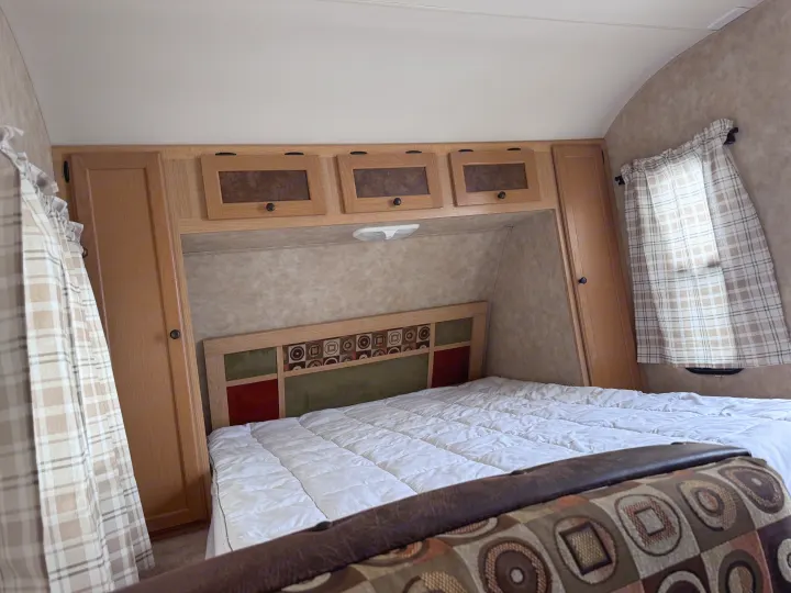 Inside an RV rental, this cozy bedroom offers a bed with patterned headboard, overhead cabinets, and plaid curtains.. kz Coyote 2010