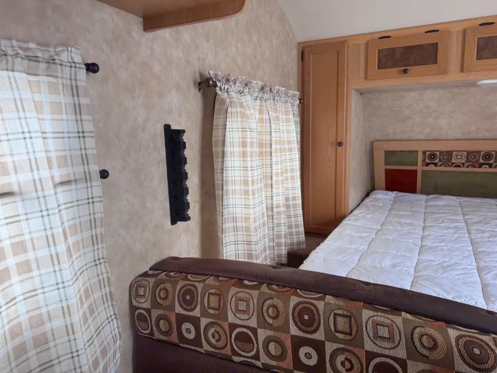 Inside a comfy RV rental: white bed, patterned headboard, plaid curtains, wooden cabinets, and geometric-print seating.. kz Coyote 2010